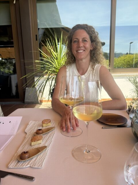 Doula agency owner smiling at an outdoor restaurant table in Spain with wine and dessert, enjoying a worry-free family vacation.